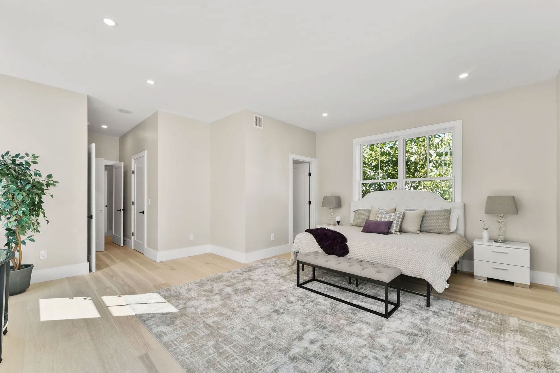 Spacious bedroom with a bed, rug, bench, and window overlooking trees. Light walls and wooden floors.