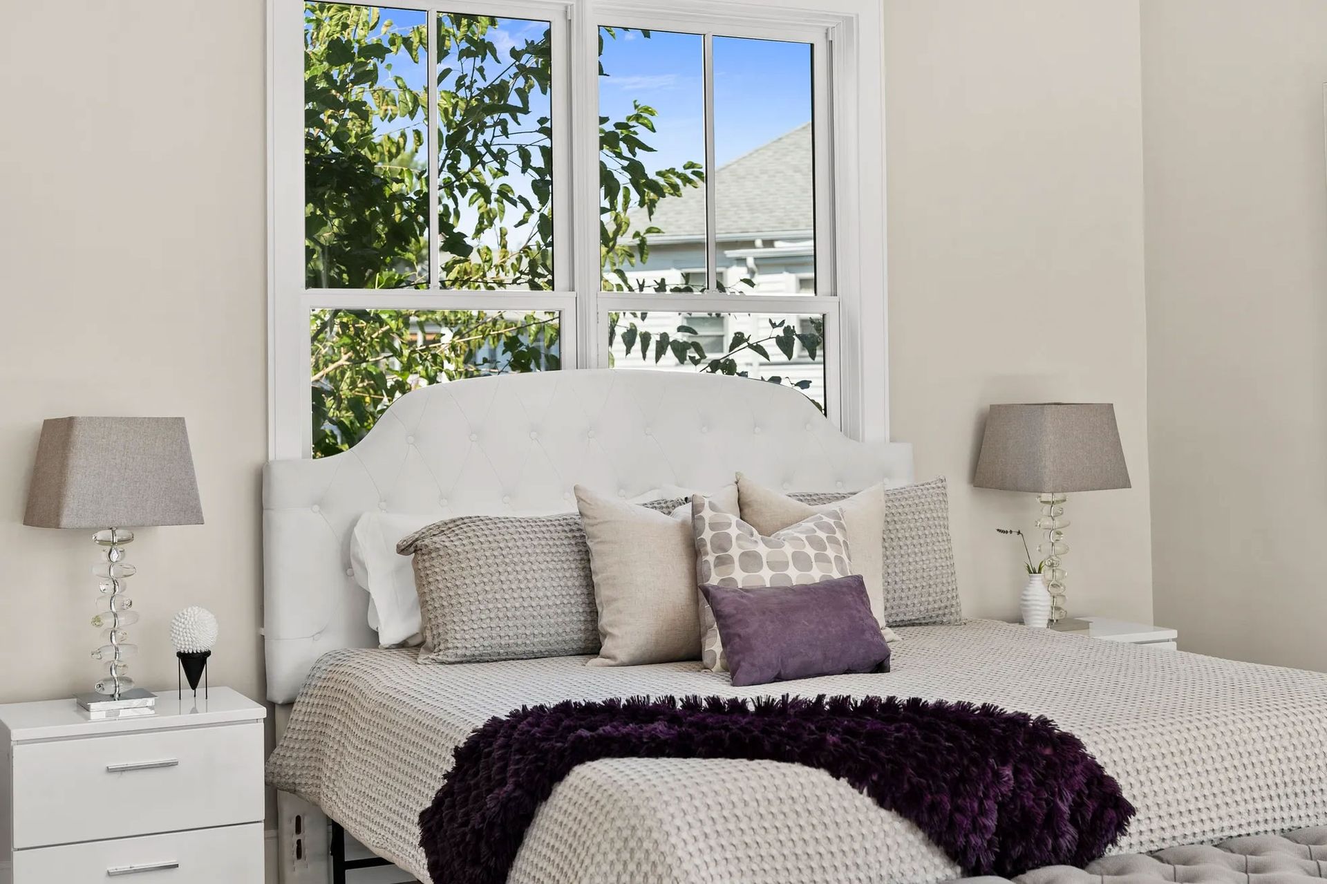 Bedroom with a white bed, neutral pillows, and purple throw blanket. Window shows trees and a building.