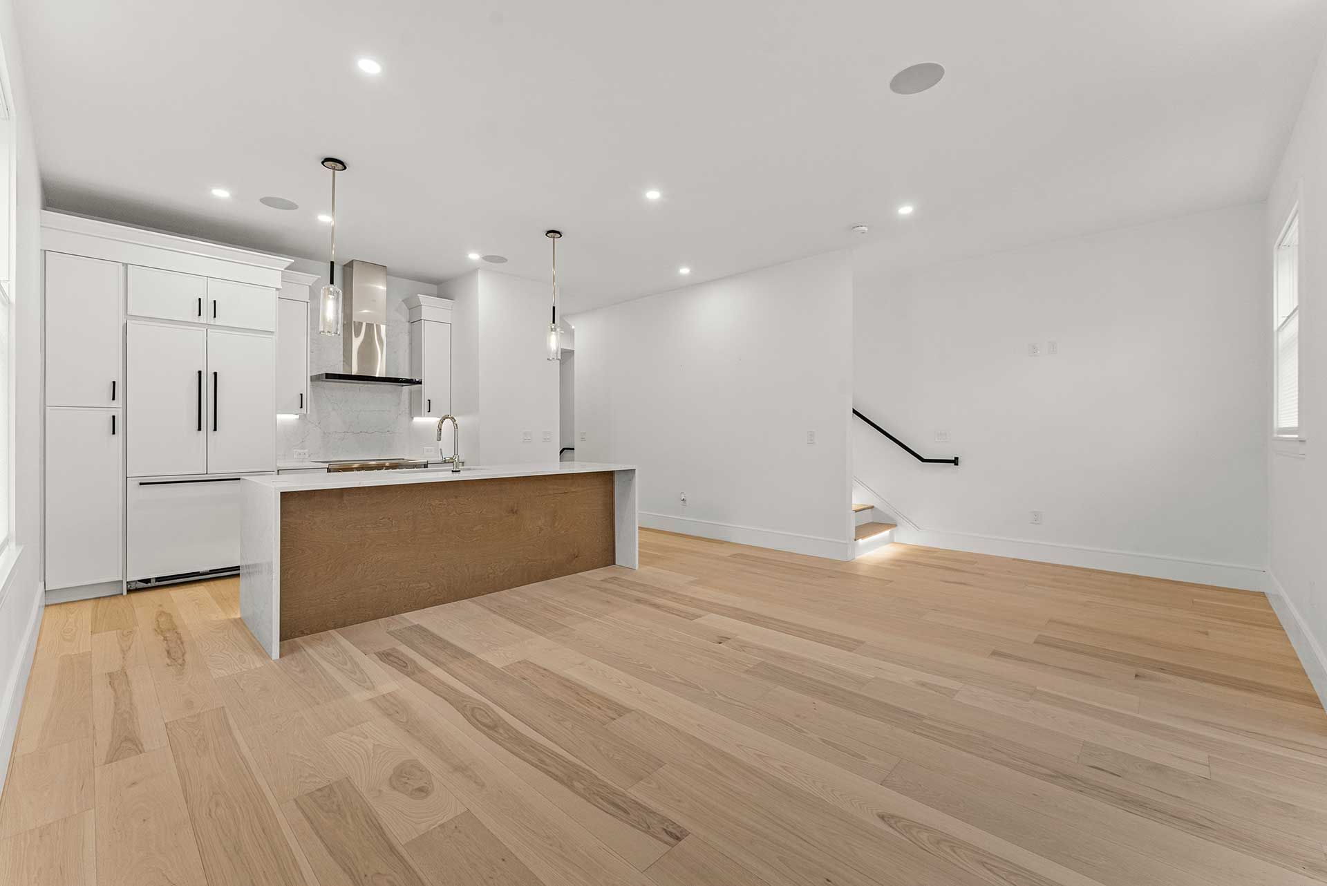 Empty, modern kitchen with white cabinetry and island, light wood flooring, and stairs.