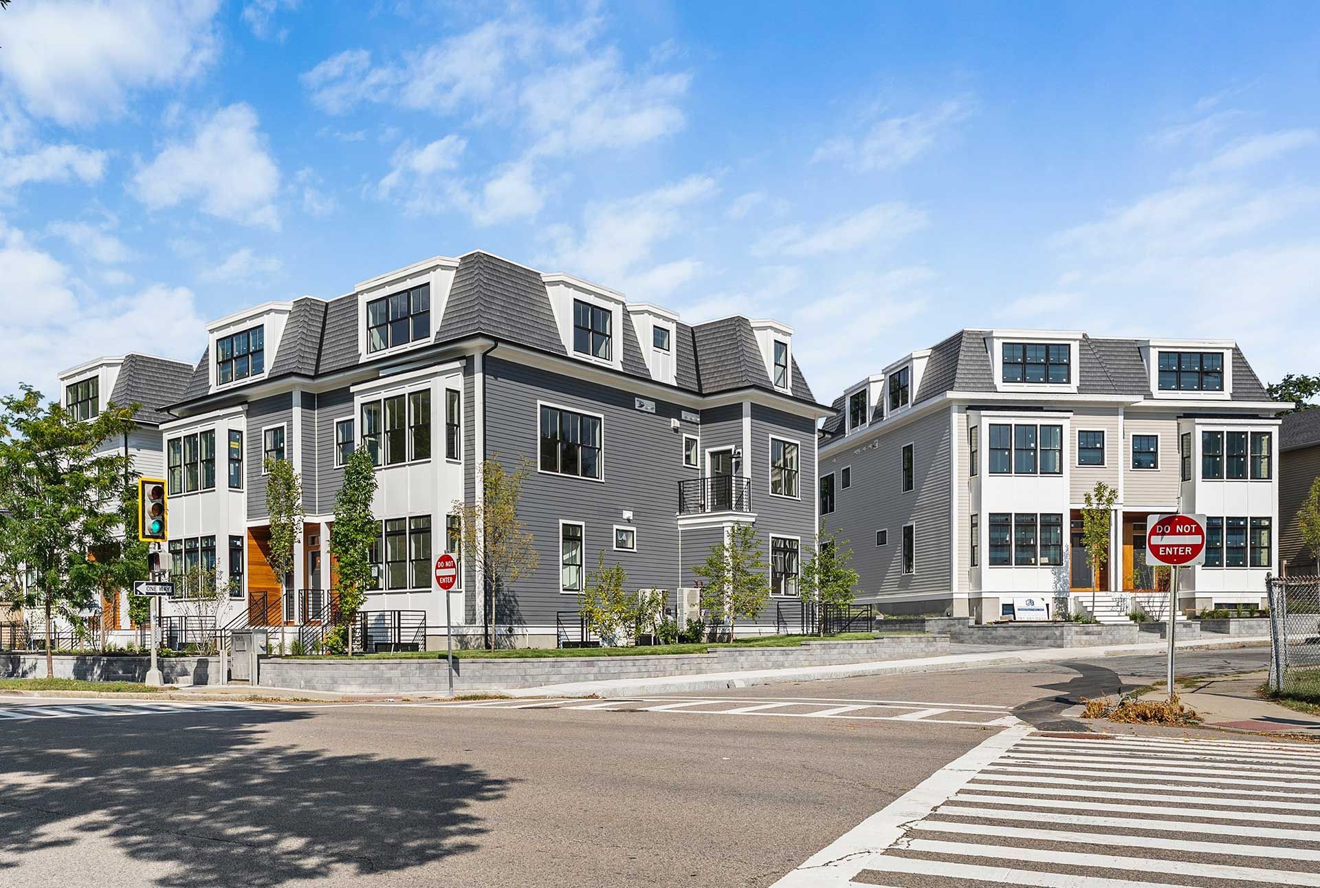 Modern gray and white townhouses with dark roofs, on a corner with crosswalks, under a blue sky.