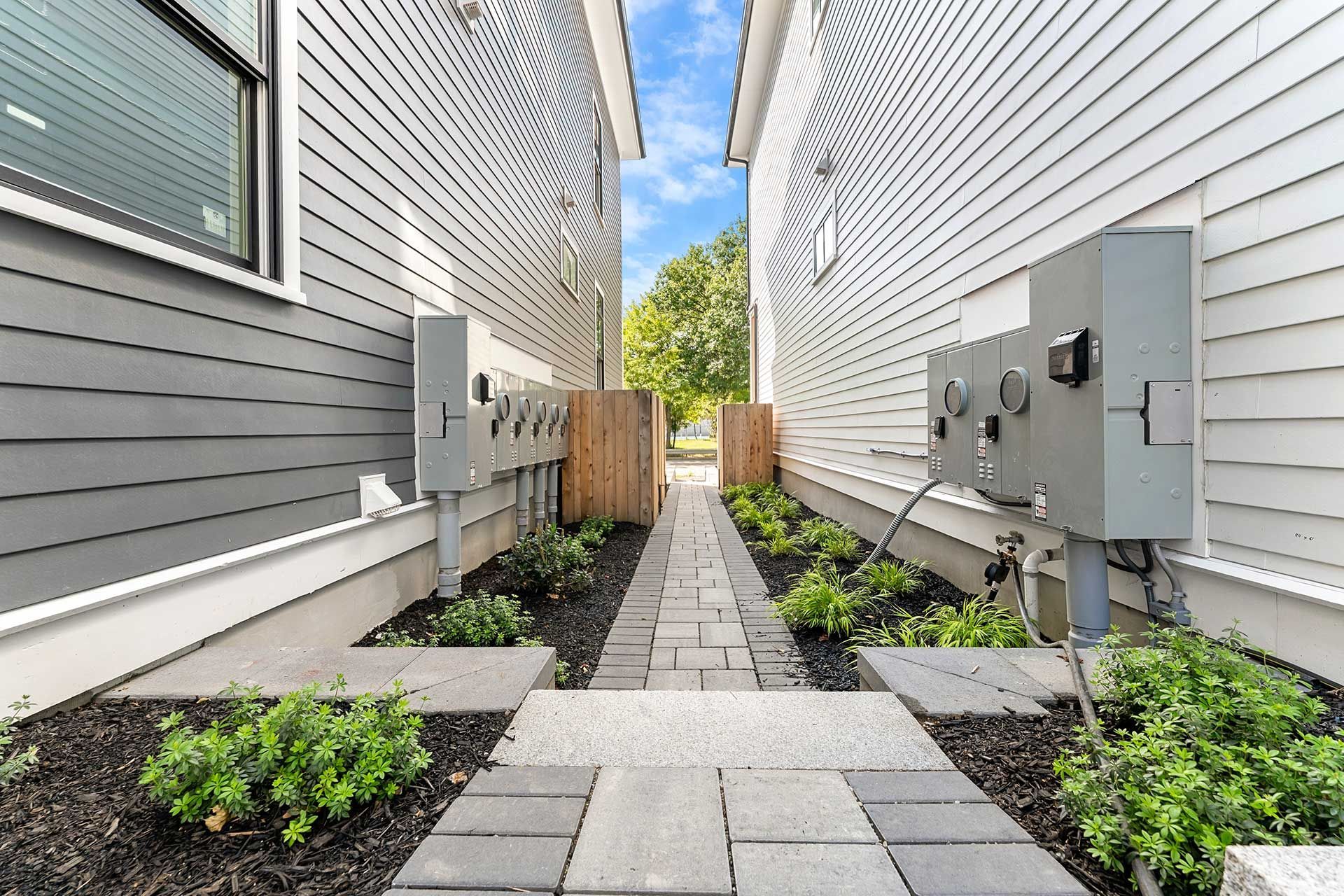 Narrow walkway between two gray-sided buildings, paved with stone and lined with bushes.