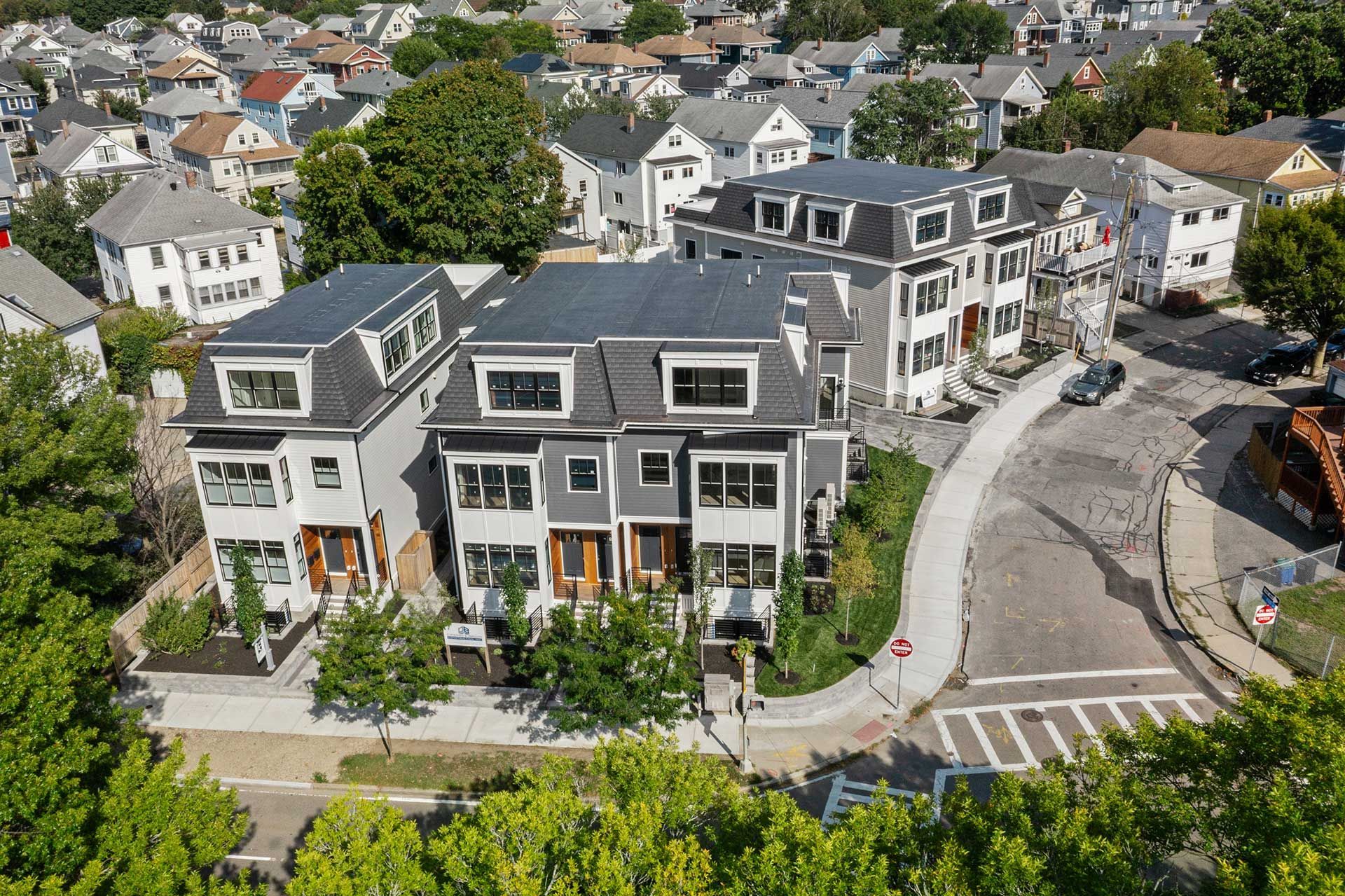Aerial view of modern grey townhouses with dark roofs on a curved street, surrounded by trees and other houses.