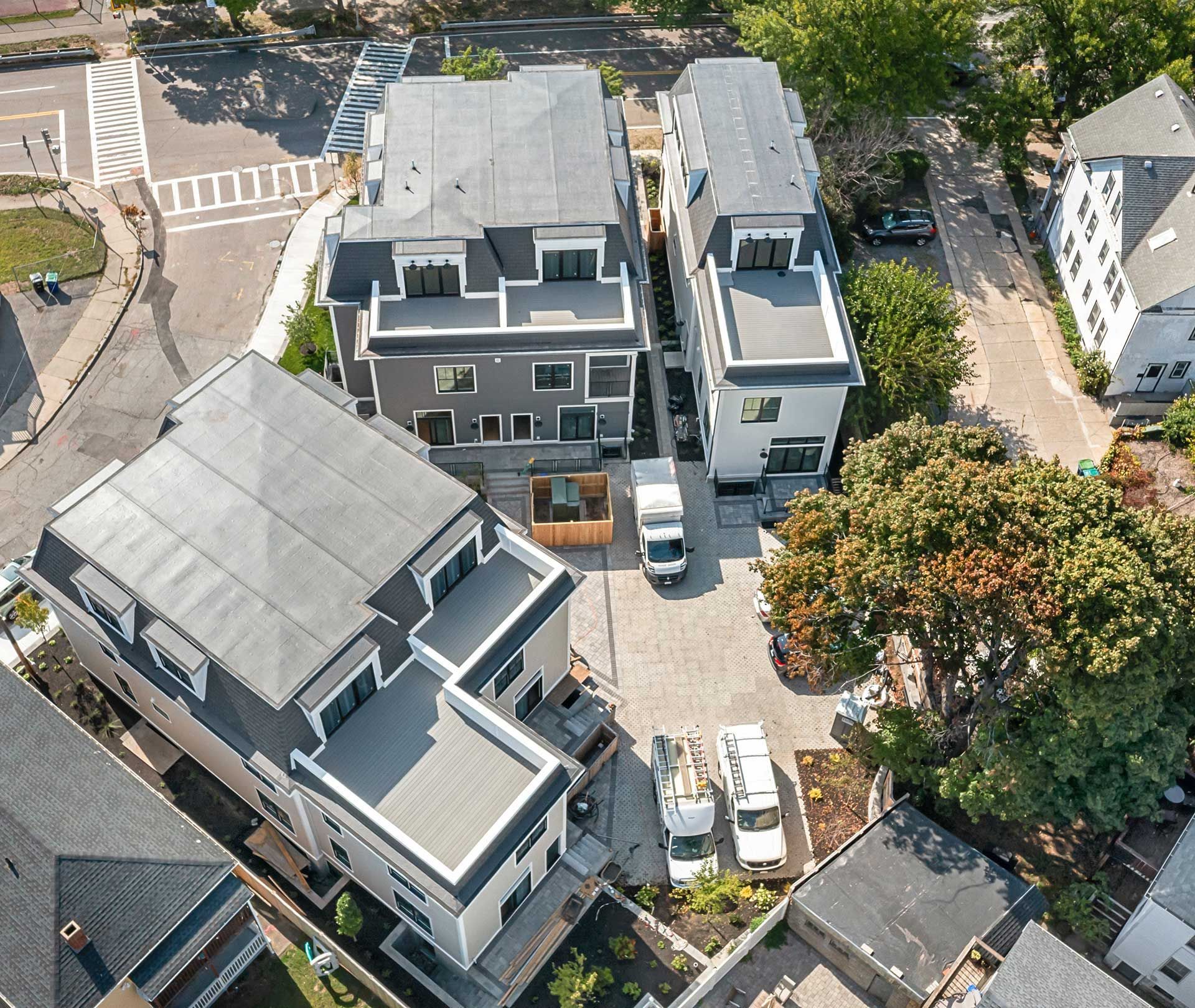 Aerial view of modern grey townhouses with flat roofs, parking lot, and surrounding trees.