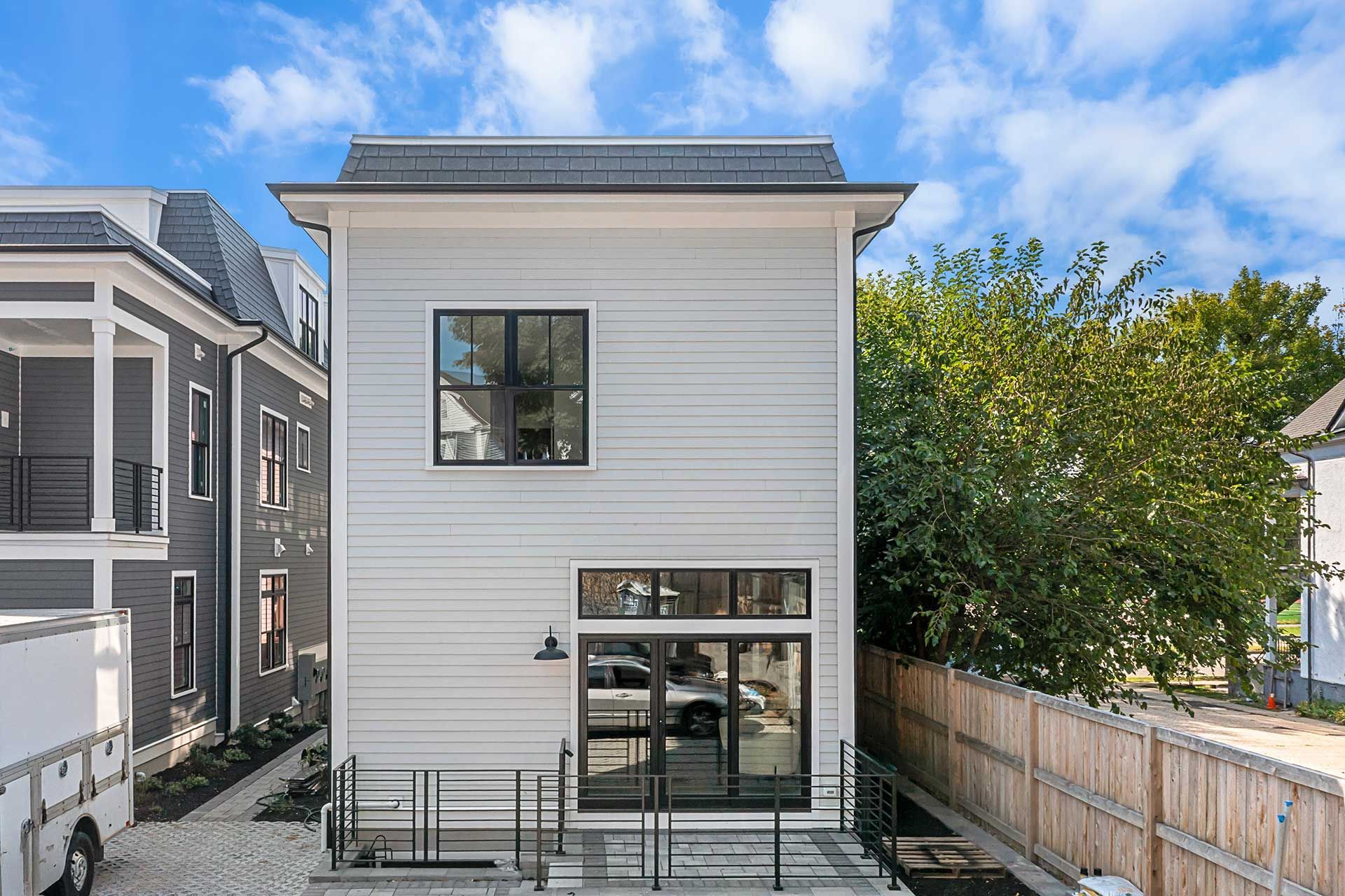 Two-story light gray house with black-framed windows, a black door, and a small wrought iron balcony under a blue sky.