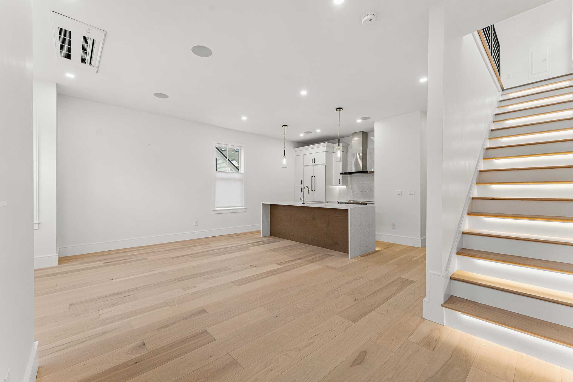 Empty, modern, all-white home interior with wood floors, kitchen island, and staircase.