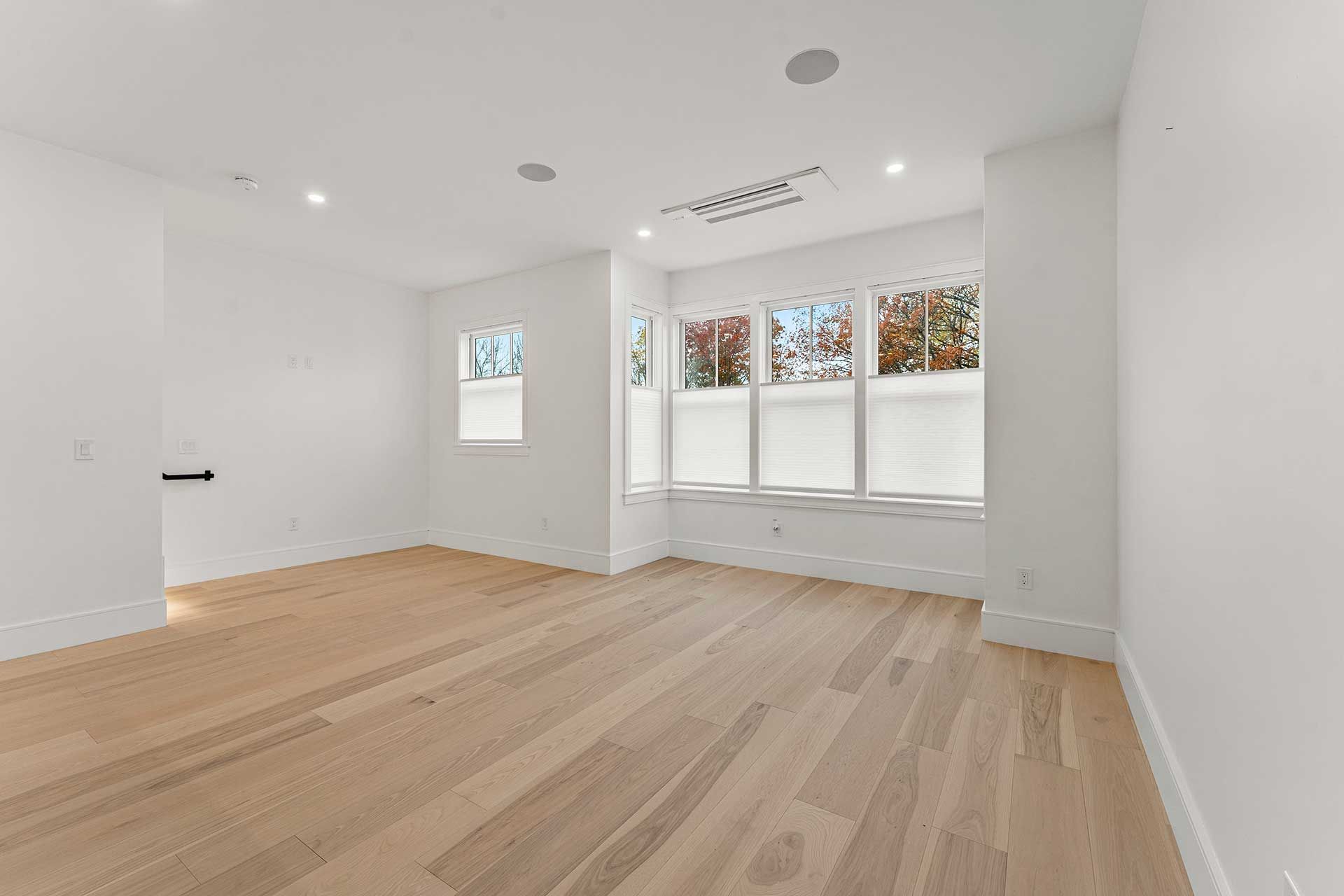Empty, bright room with hardwood floors, white walls, and multiple windows with blinds.
