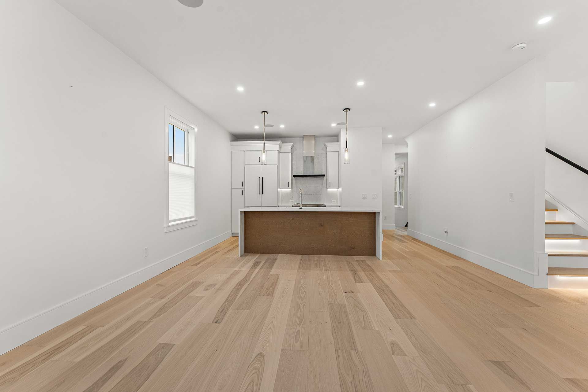 Empty, modern open-plan living space with kitchen island and stairs. Light wood floors, white walls, and recessed lighting.