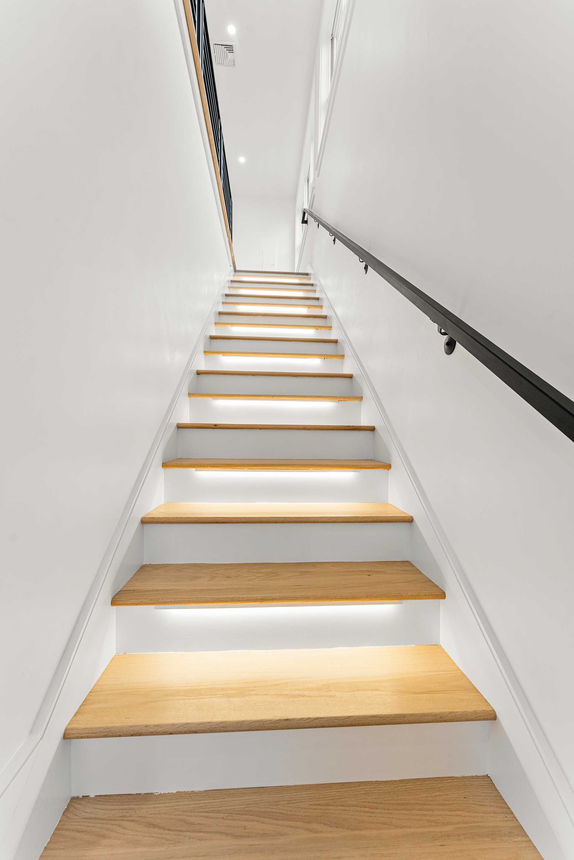 Wooden stairs with under-step lighting ascend between white walls, a black handrail on the right.