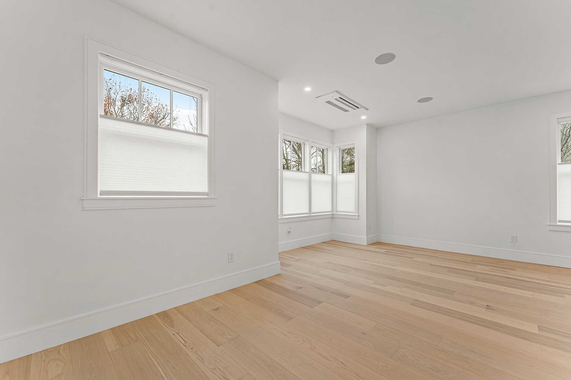 Empty, white room with light wood floors, three windows with shades, and recessed lighting.