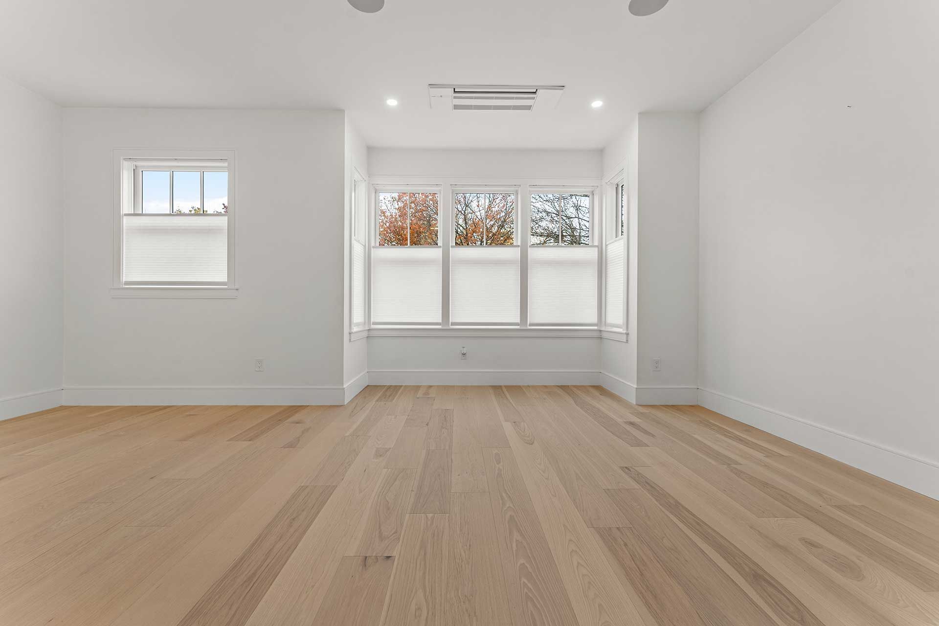 Empty room with light wood floors, white walls, and three windows with neutral shades.