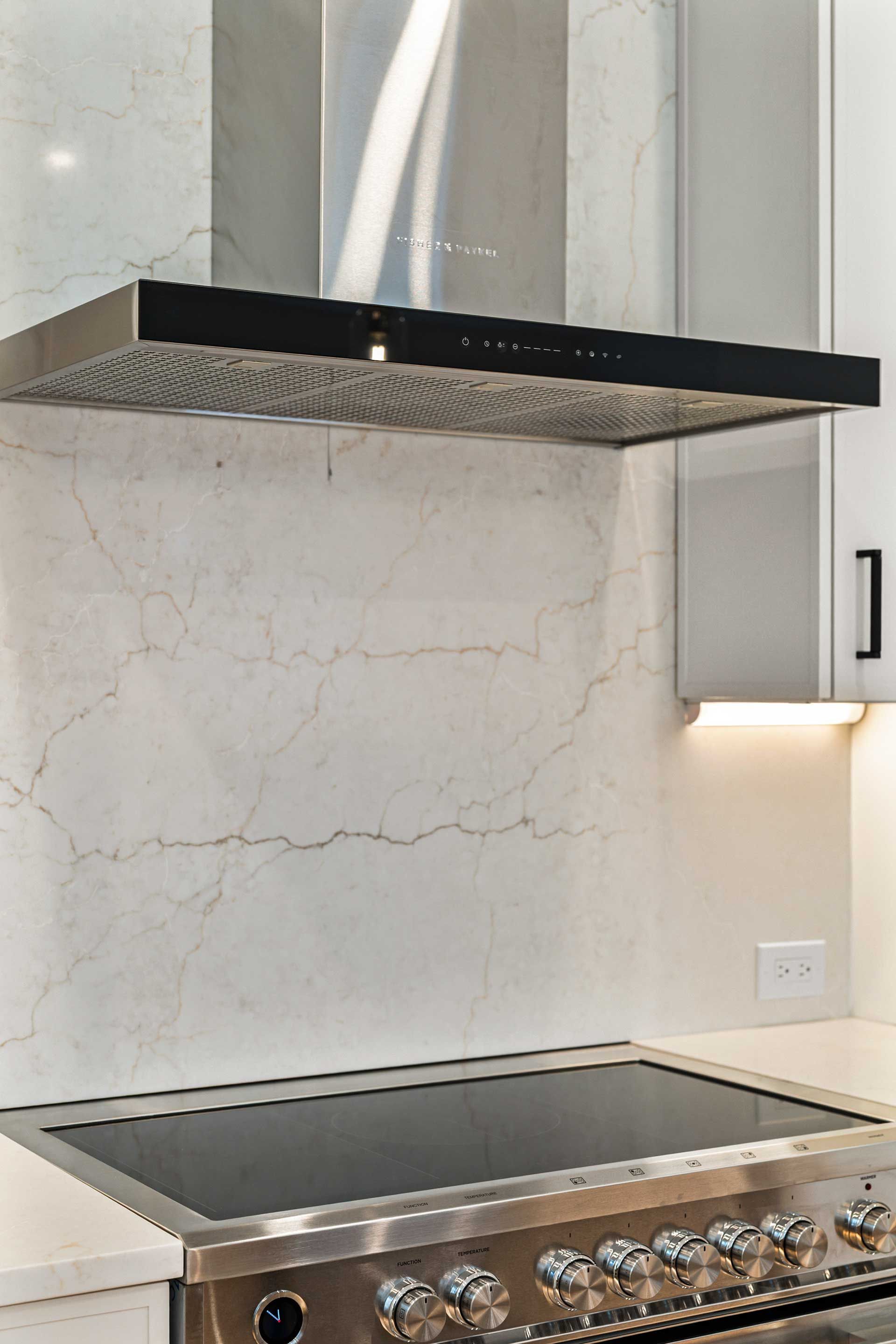 Stainless steel range with black cooktop, under a range hood in a white kitchen with marble backsplash.