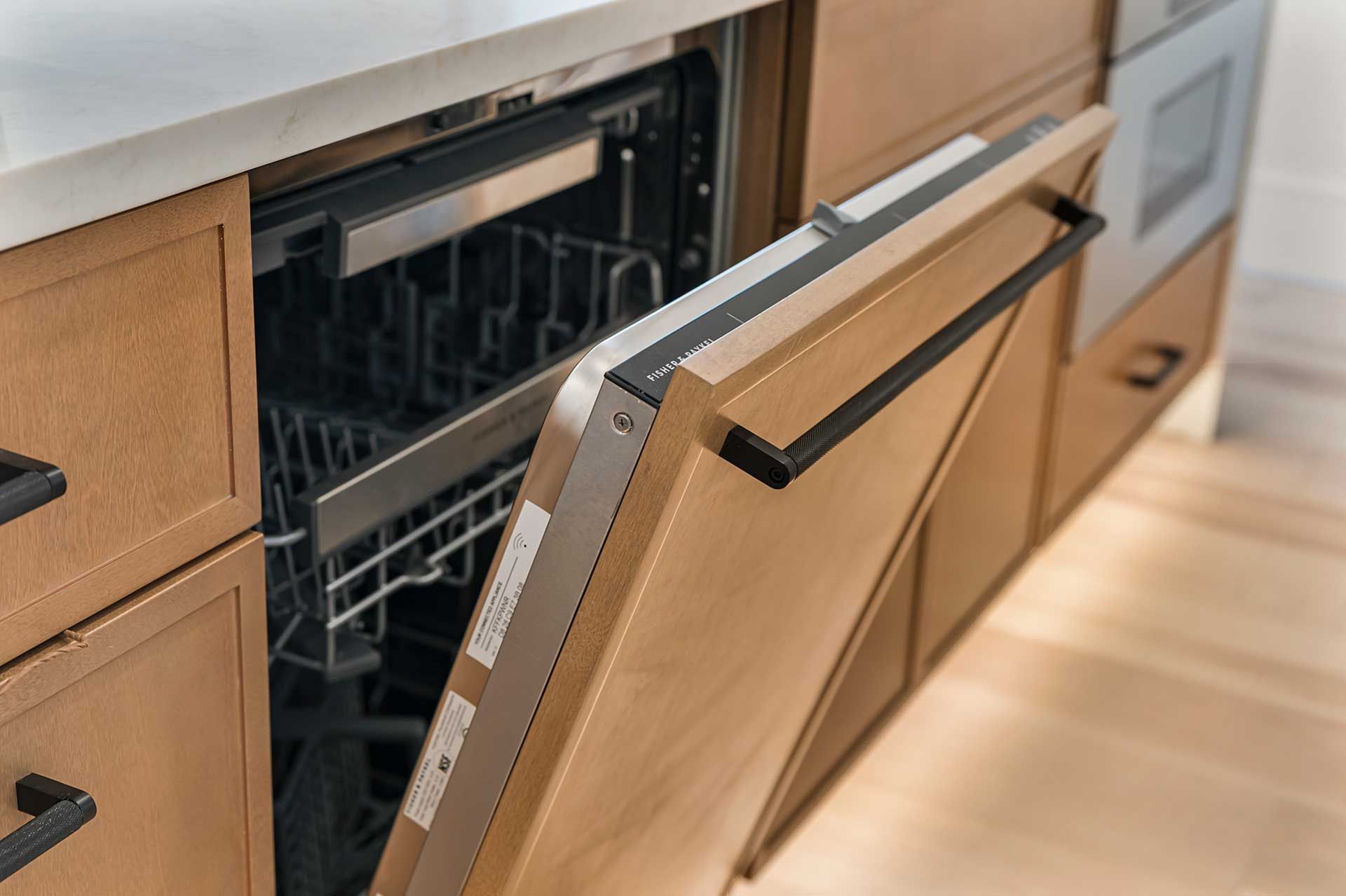 Open dishwasher in a light brown wooden cabinet. Stainless steel interior, black handle.