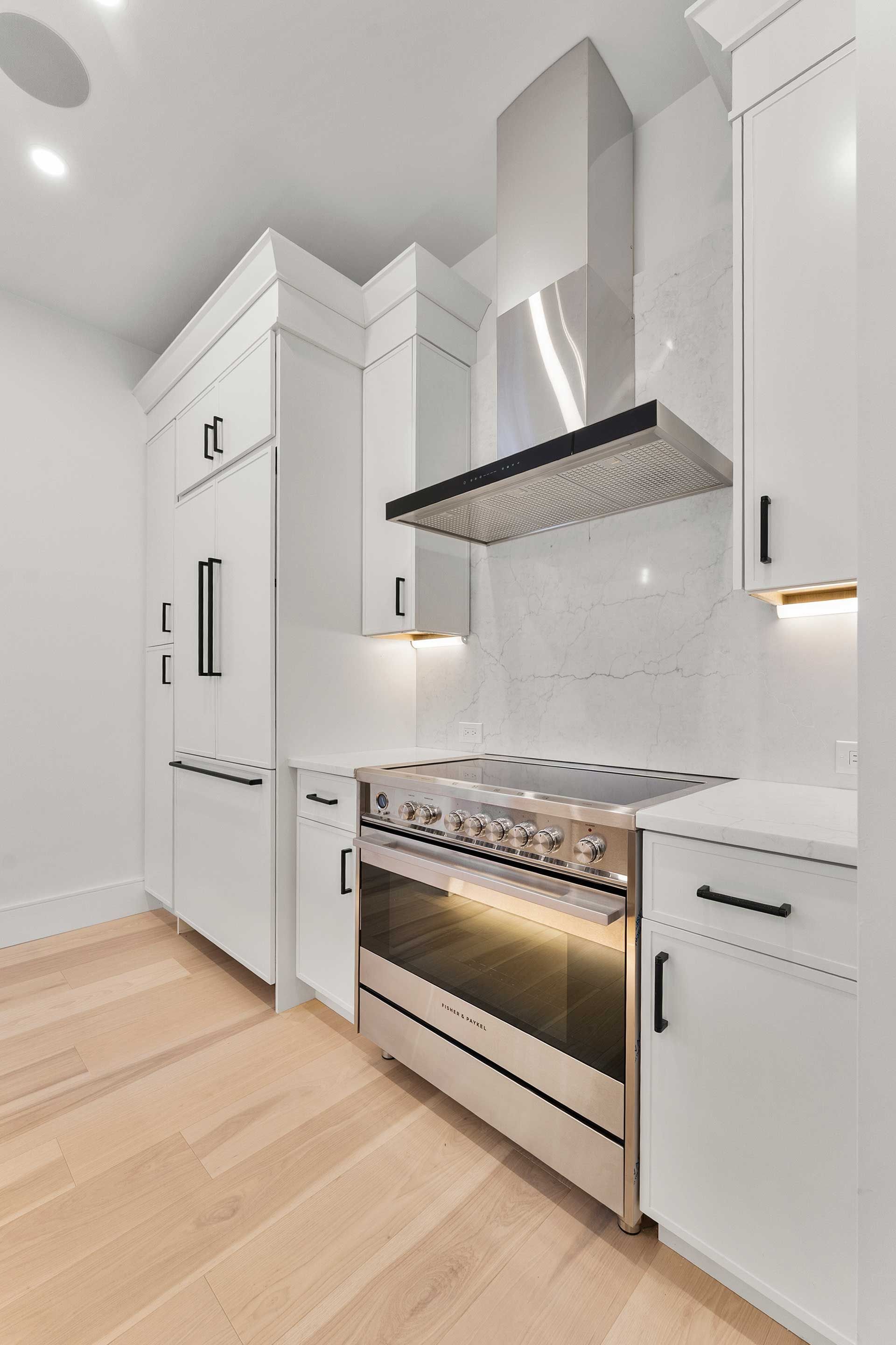 Modern white kitchen with stainless steel oven, hood, and light wood flooring.