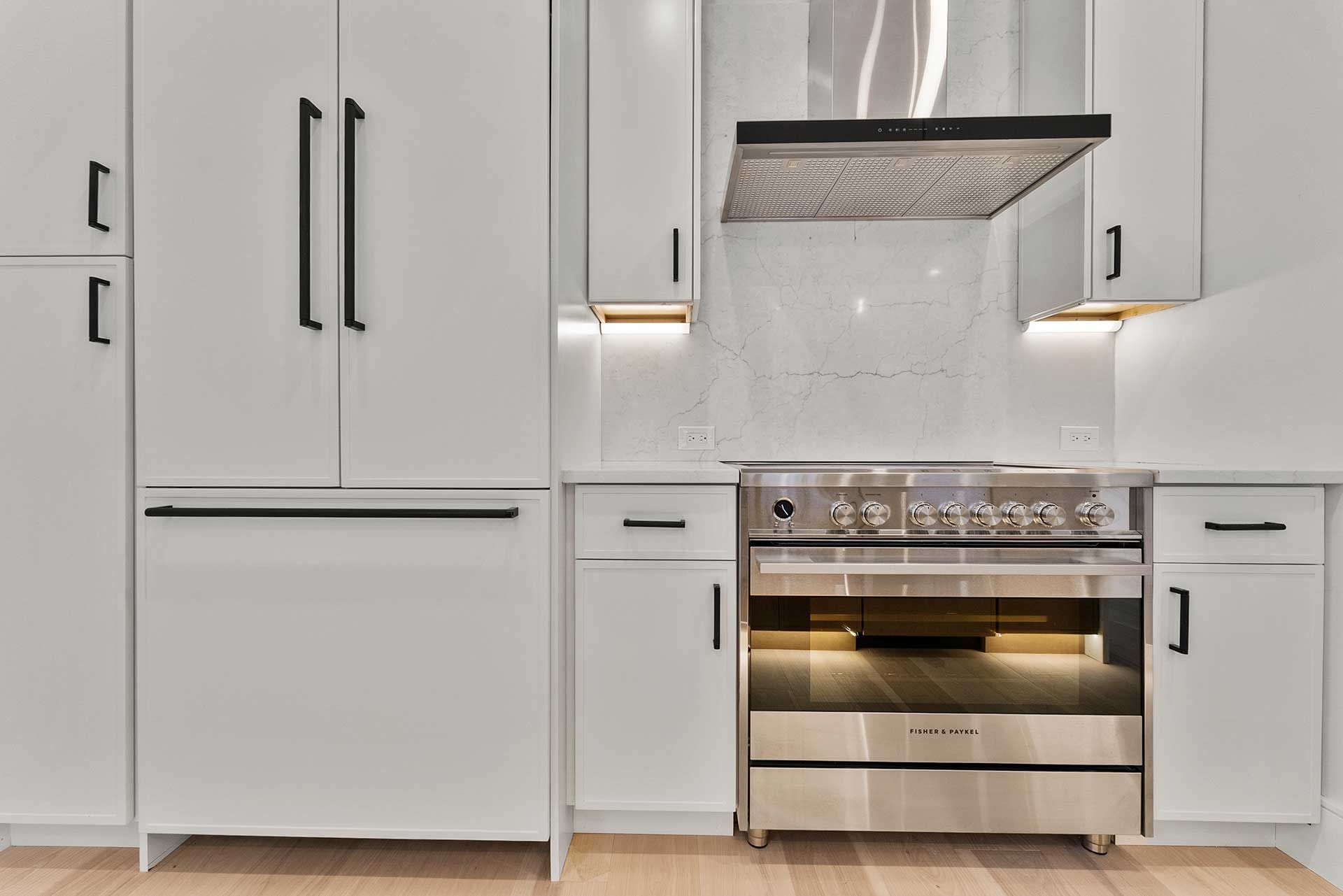 White kitchen with stainless steel oven and range hood. Cabinets have black handles, set against white speckled backsplash.