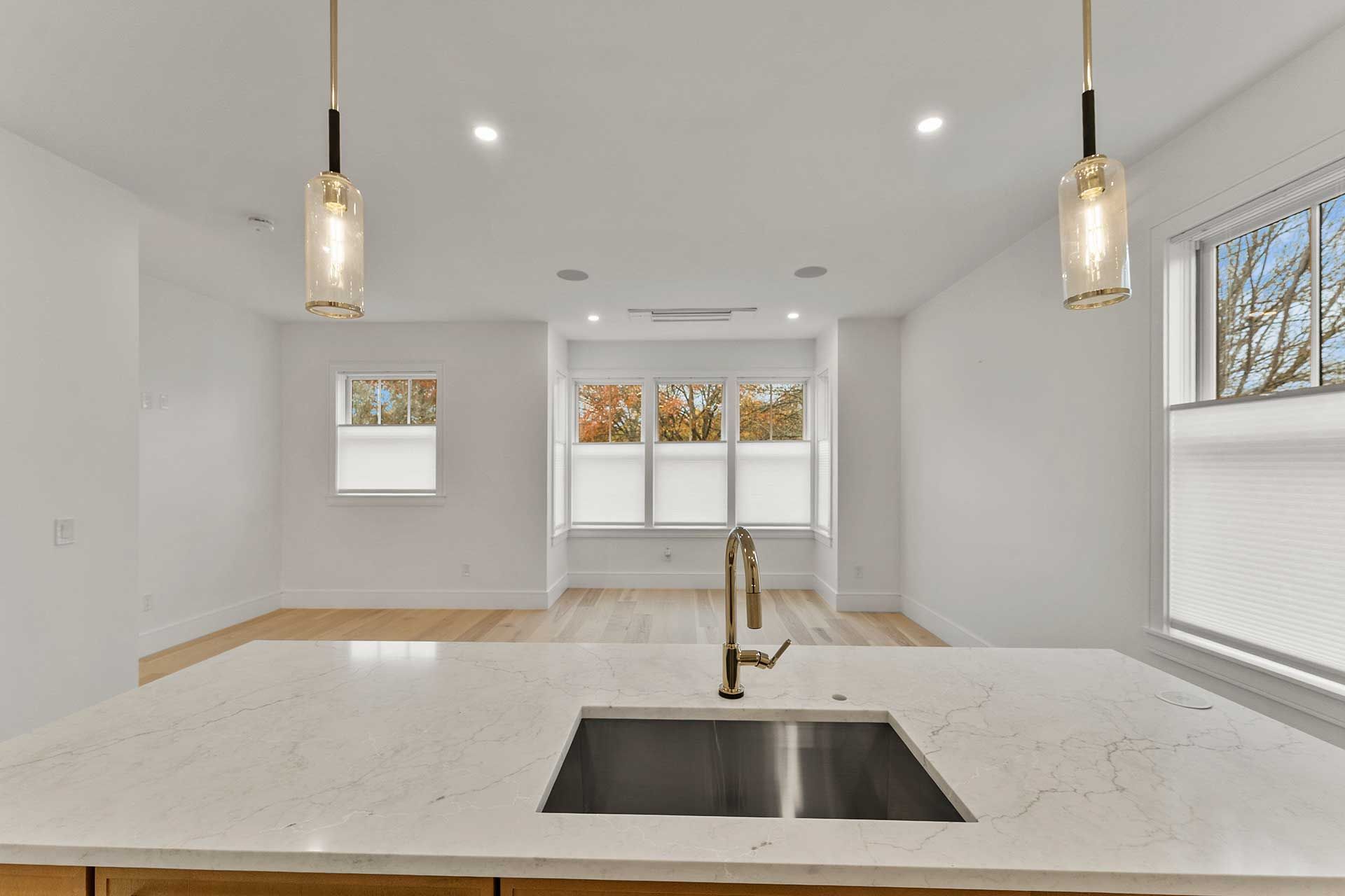 Modern kitchen with light wood floor, white countertops, and three windows overlooking trees.