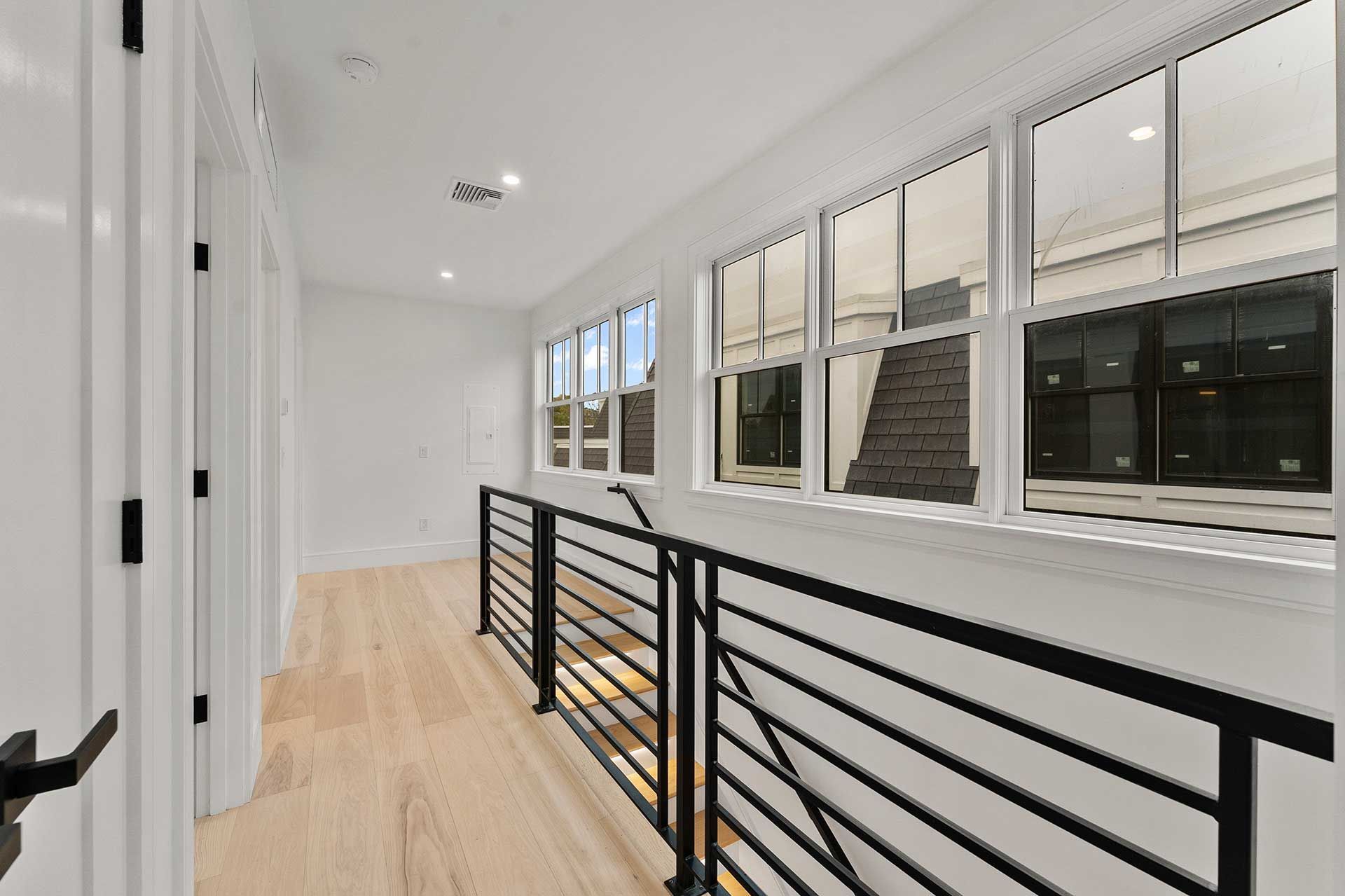 Hallway with light wood floors, white walls, black railing, and windows overlooking a stairwell.