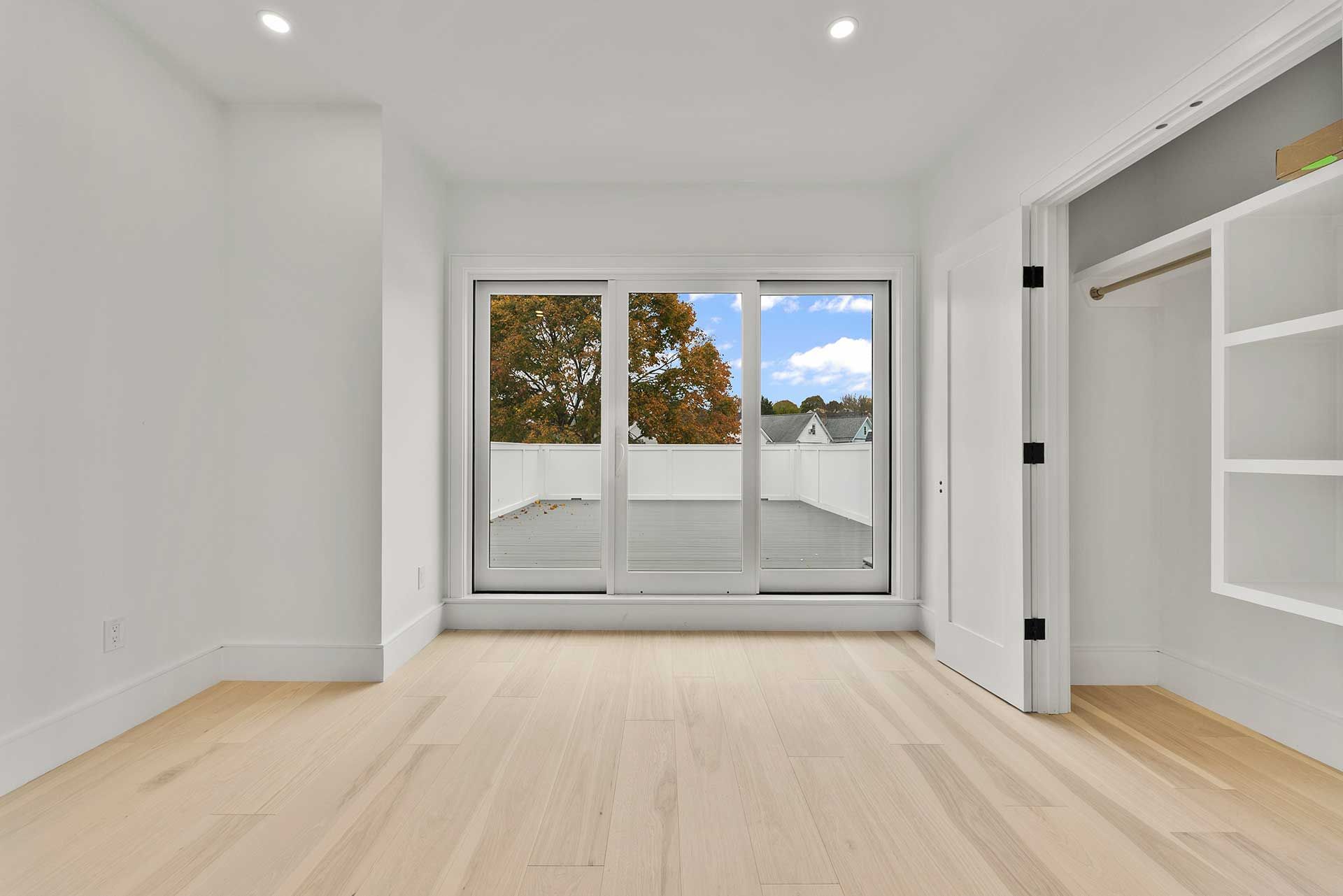 Empty, bright bedroom with large window overlooking a balcony.  Wooden floor, white walls, and built-in closet.