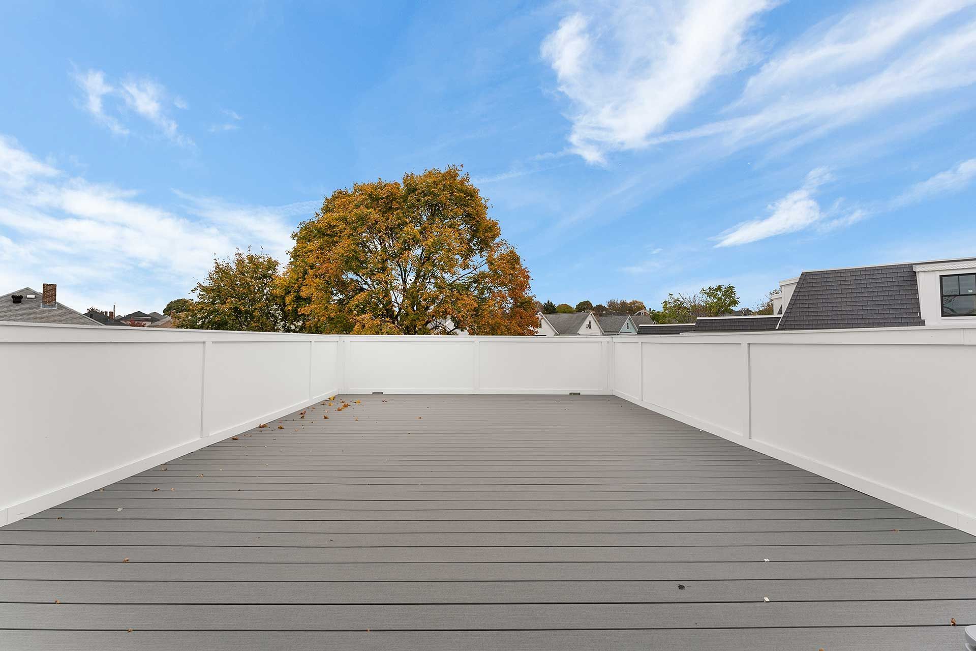 Gray wooden deck with white railing, blue sky and autumn tree in the background.