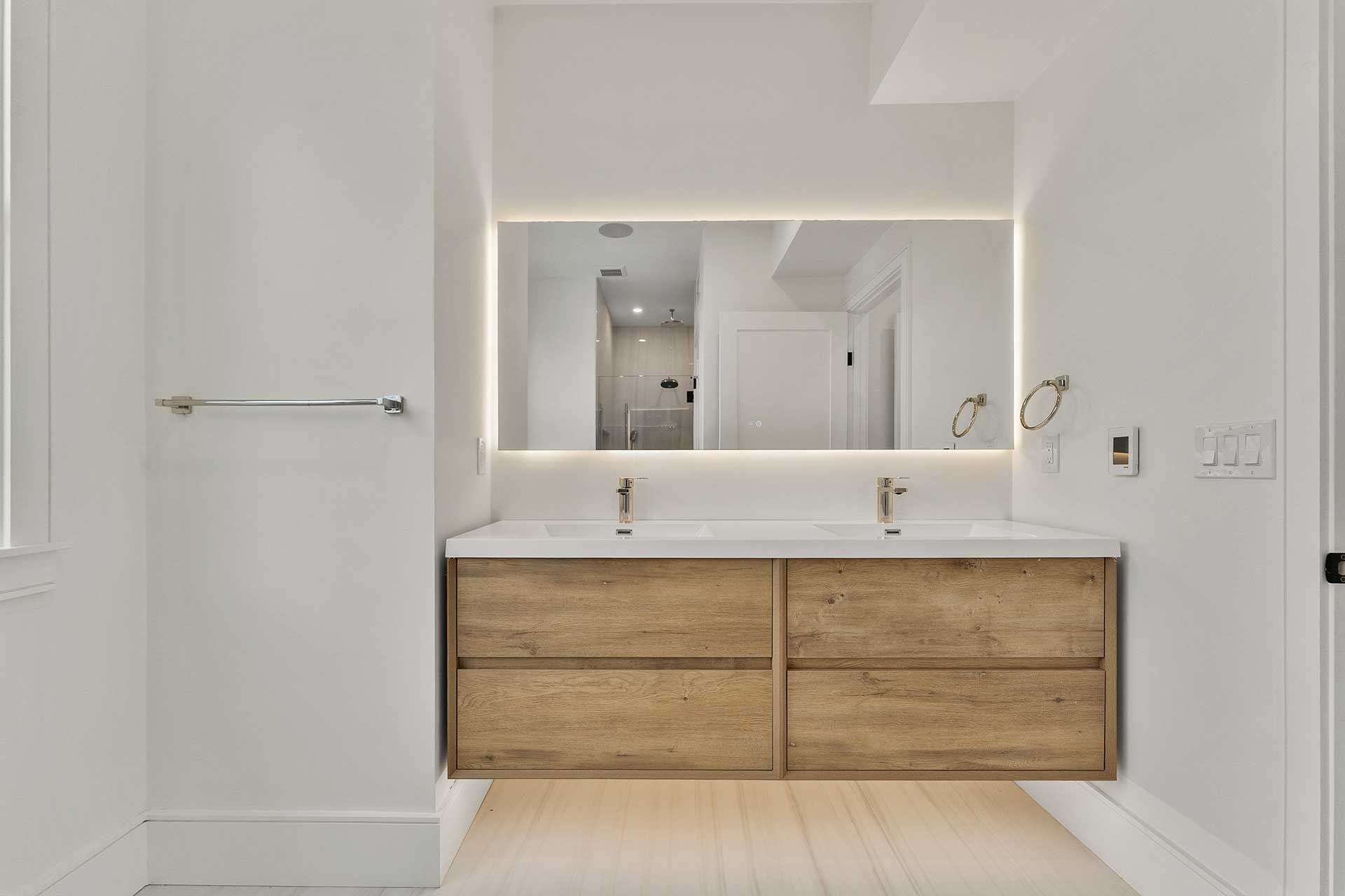 Modern bathroom with floating wooden vanity, large mirror with backlighting, and two sinks.