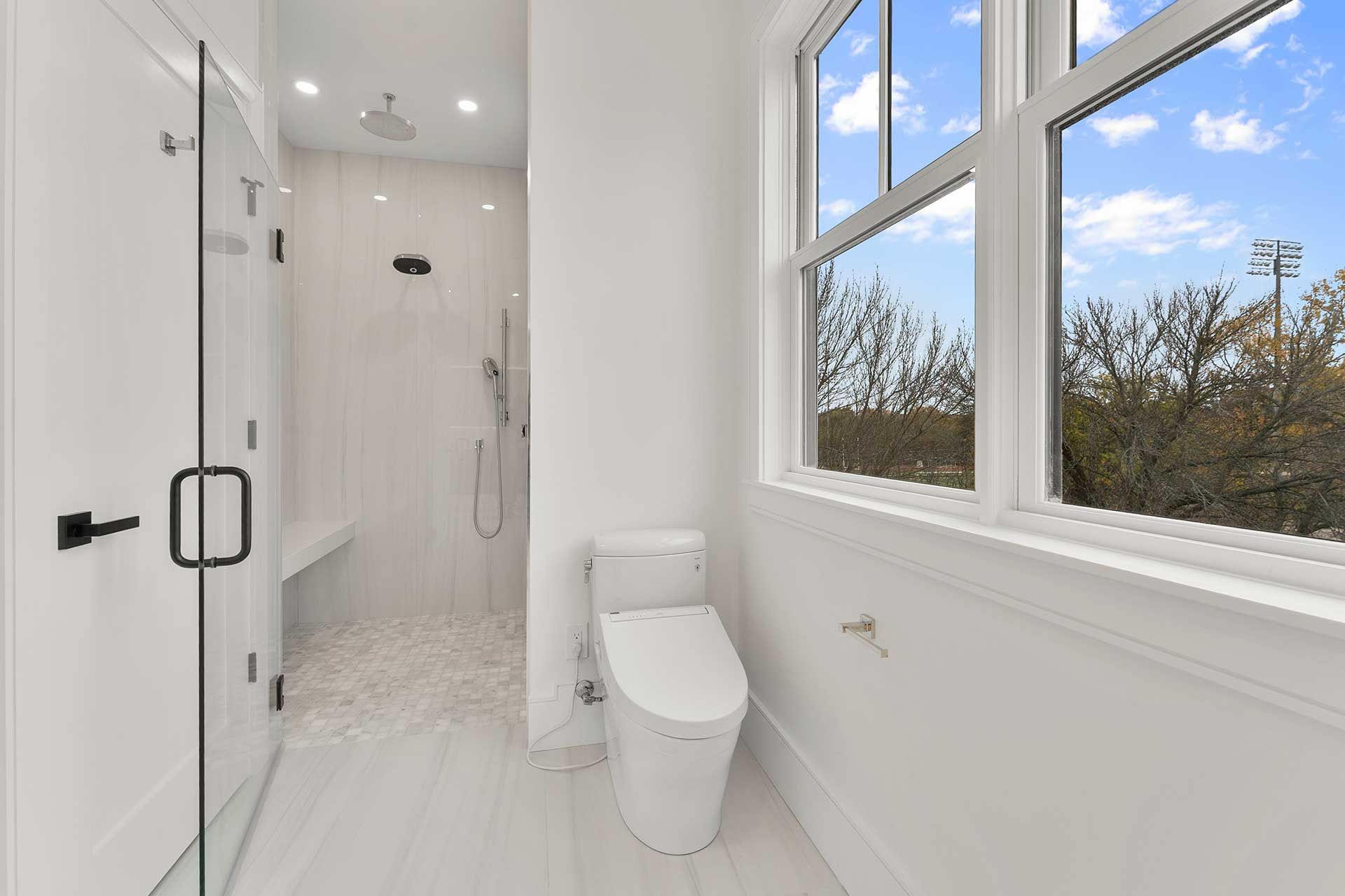 Bright white bathroom with a glass shower, toilet, and a window with an outdoor view.
