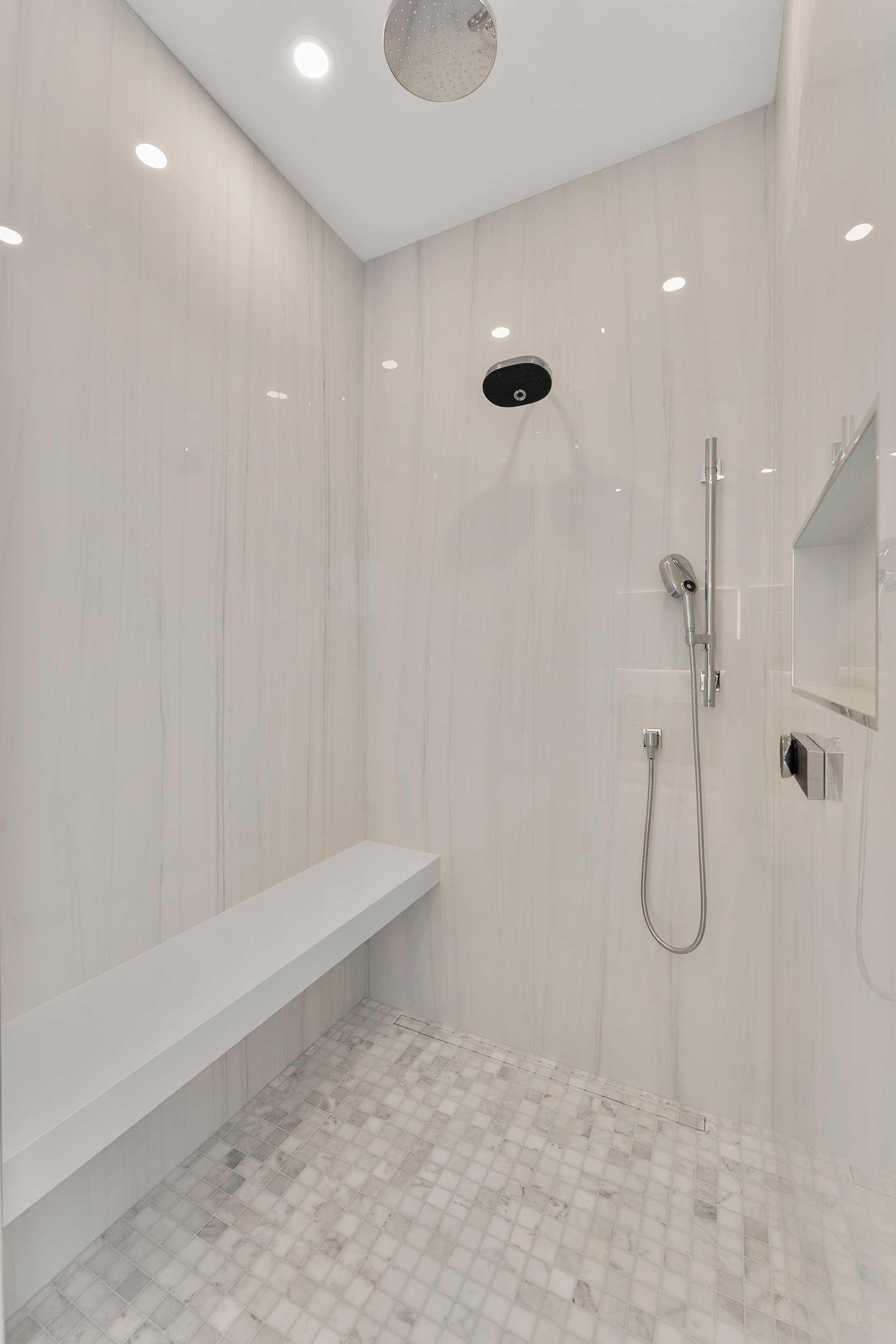 White-tiled shower with built-in bench, overhead and handheld showerheads, and small mosaic floor tiles.