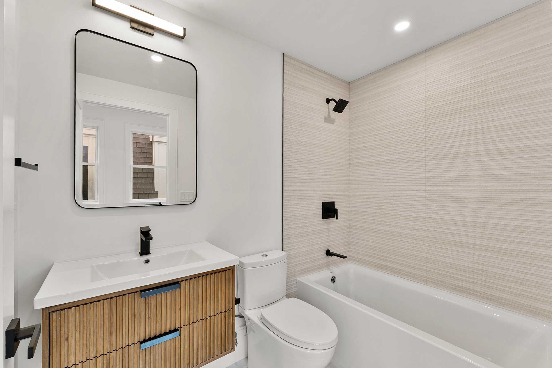 Bathroom with a wooden vanity, a white toilet, and a bathtub with a tiled wall. Black fixtures and a mirror.
