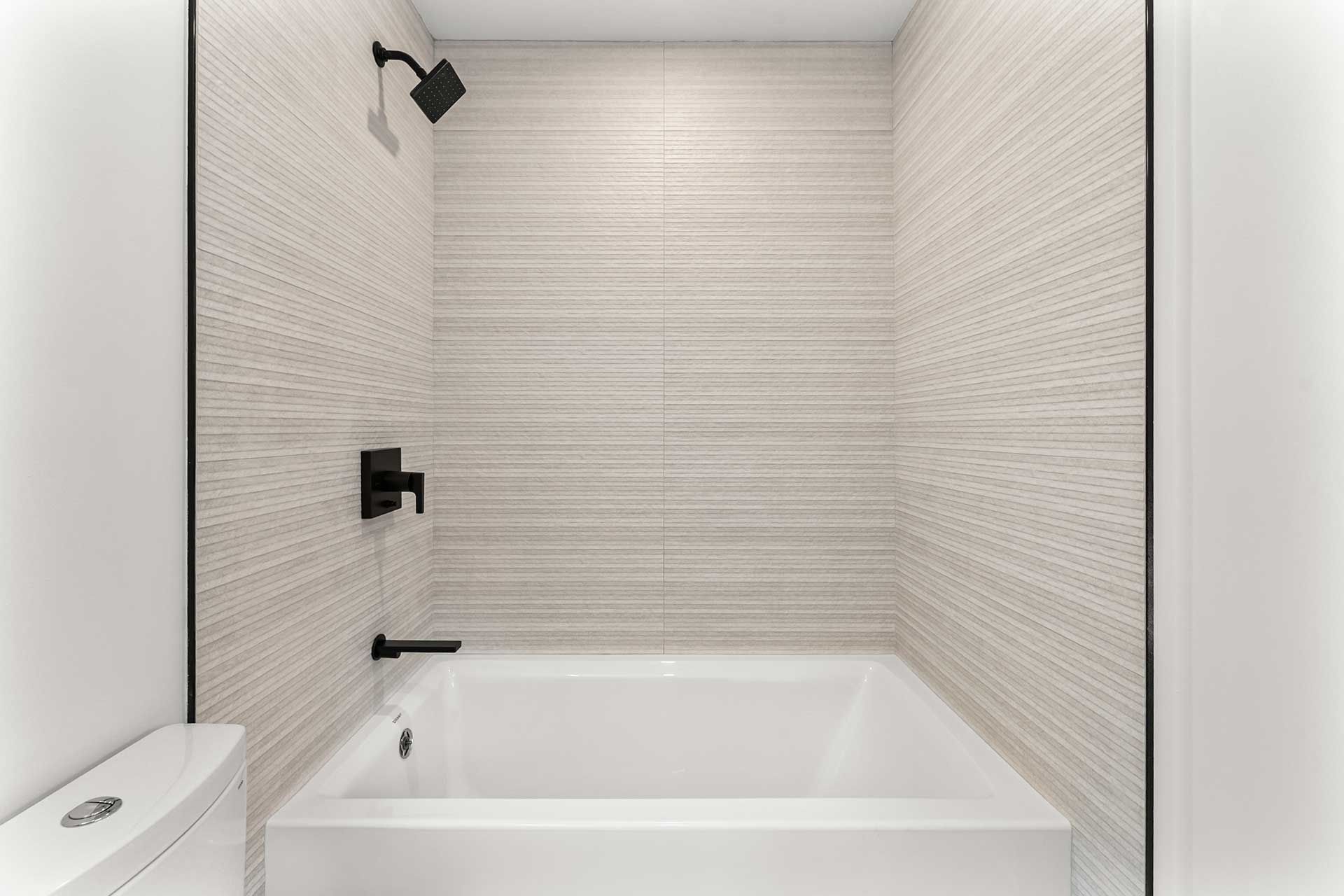 Small bathroom with white tub, showerhead, and textured beige tile walls. Black fixtures.