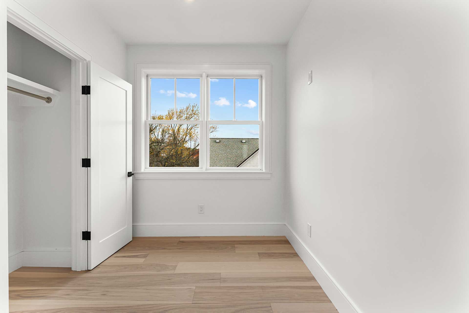 Empty, bright room with a window, wooden floor, and white walls. A door on the left is open.