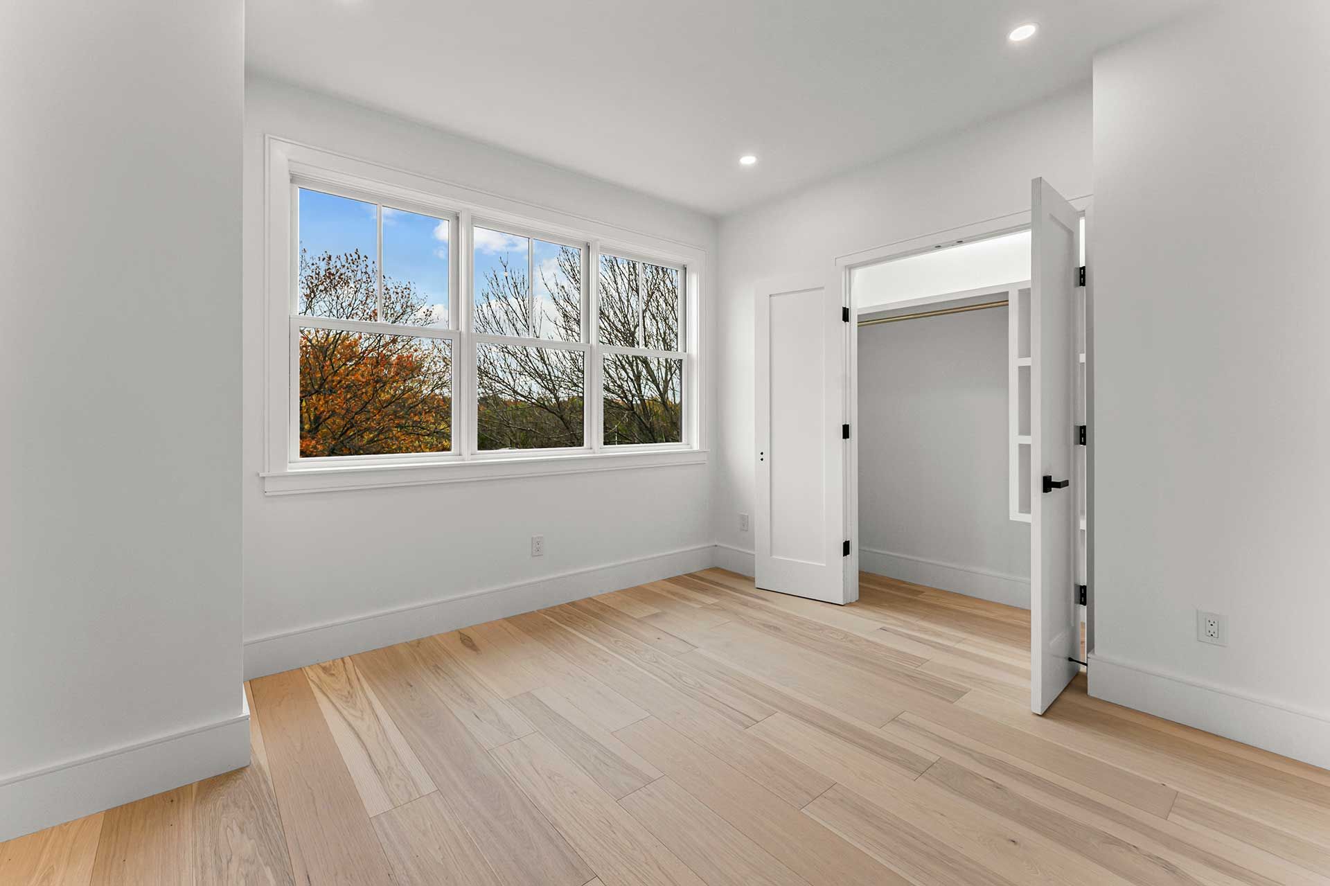 Empty, bright bedroom with wood floors, white walls, large window, and open closet door.