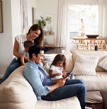 Family of four relaxing on a couch, looking at a tablet, in a bright living room.