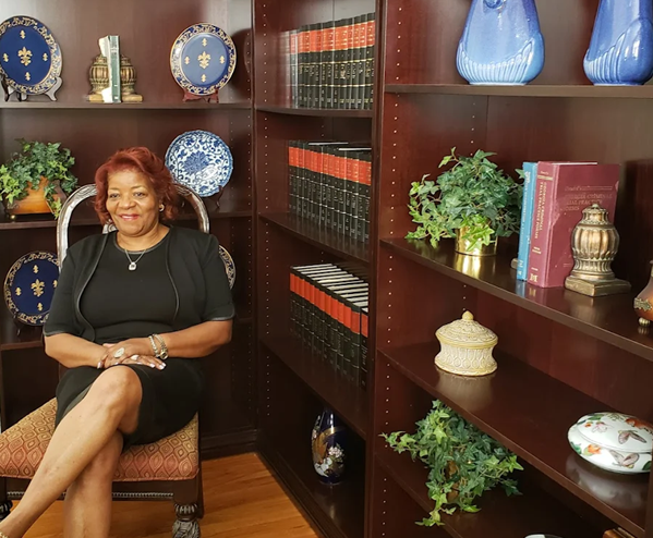 Woman seated in front of a bookshelf, smiling. Dark dress, dark hair. Books, decorative plates, and vases on the shelves.