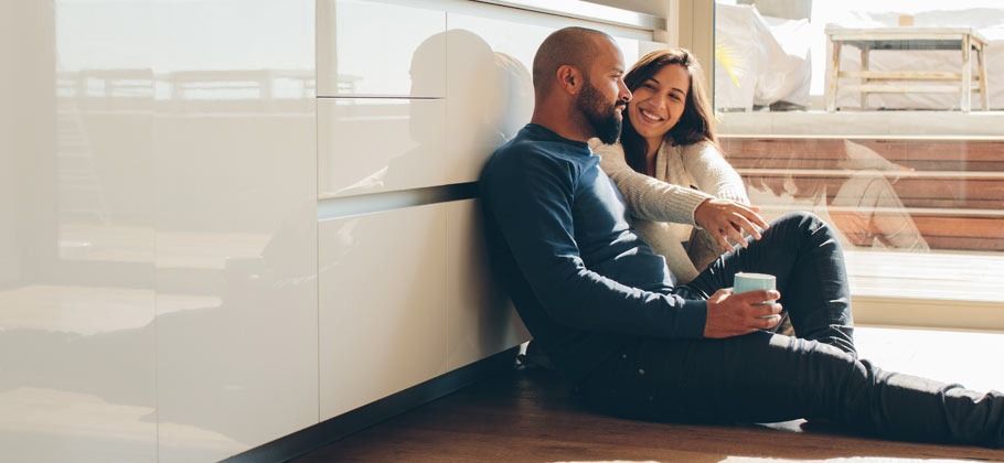 Couple sitting on floor, leaning against a white structure, looking at each other, smiling.