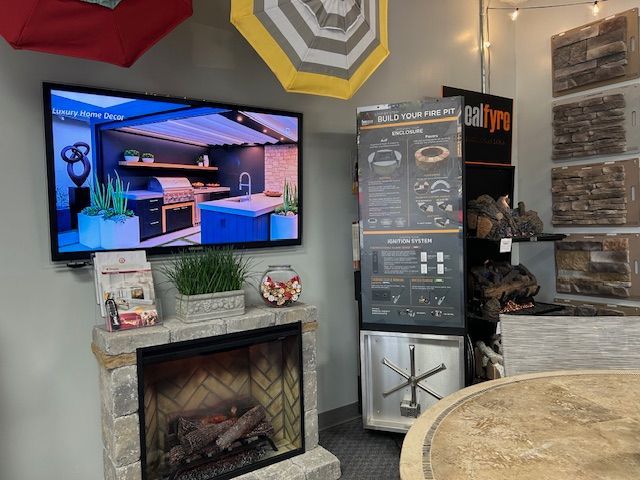 Display with a TV showing an outdoor kitchen, fireplace, product display, and patio umbrella samples.
