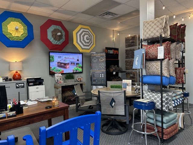 Interior retail space displaying patio furniture and accessories. Colorful umbrellas adorn the wall, pillows on shelves, desk, and chairs.