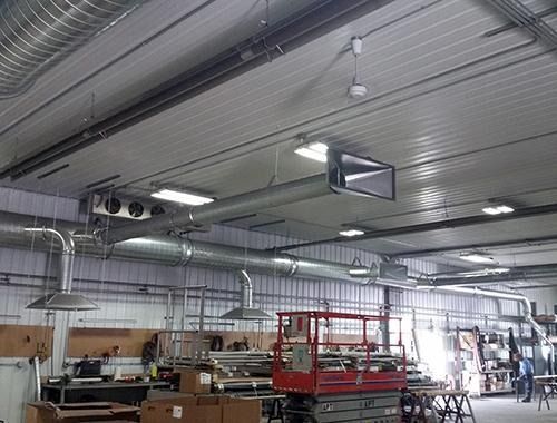 Interior of a factory workshop with metal ceiling, hanging lights, and worktables beneath a large monitor