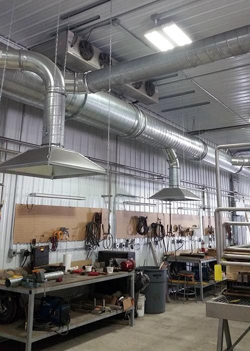 Industrial workshop with metal ductwork, hanging lights, and workbenches along the wall