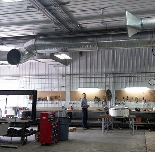 Industrial workshop with metal ductwork, workbenches, and a person standing in the center