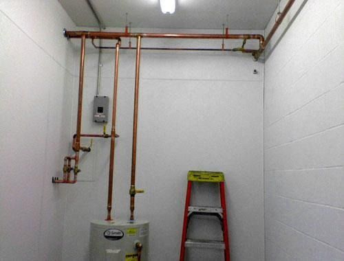Utility room with copper pipes, water heater, and a red ladder against white tiled walls.