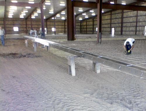 Two workers inspecting a long metal beam inside a large warehouse with a dirt floor