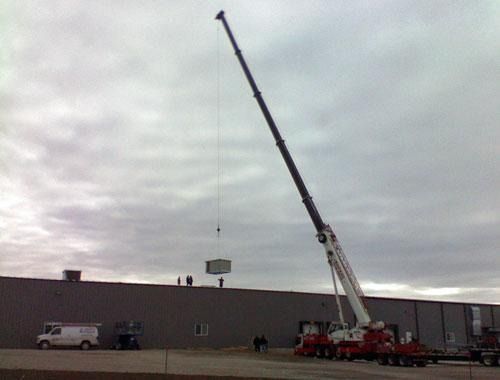 Crane with extended boom beside a warehouse under a cloudy sky, with a white van nearby.