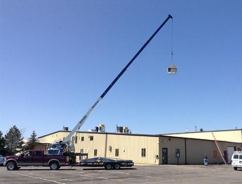 Crane lifting a small basket beside a tan warehouse, with a red truck and trailer in the lot