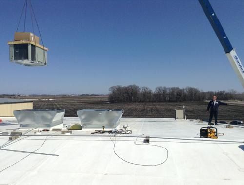 Crane lifting a rooftop HVAC unit onto a white roof with workers nearby under a clear blue sky