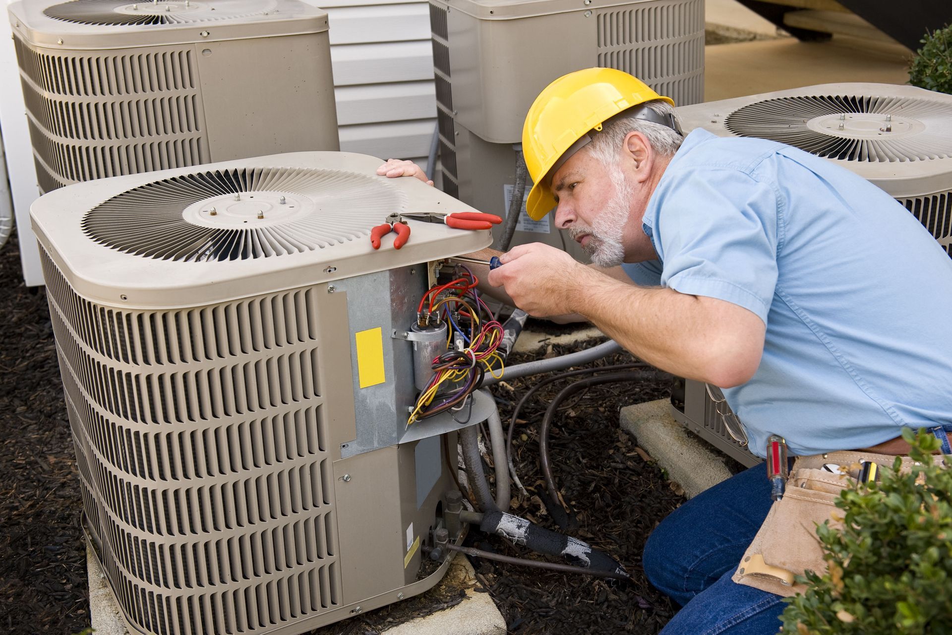 Technician in yellow hard hat repairing outdoor HVAC unit with exposed wiring