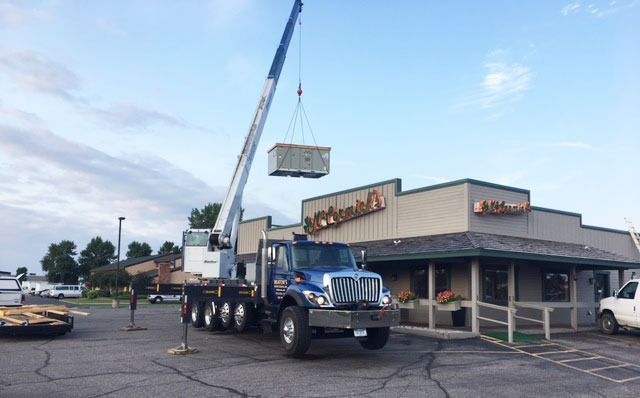 Crane lifting a rectangular sign onto a truck parked outside a commercial building.