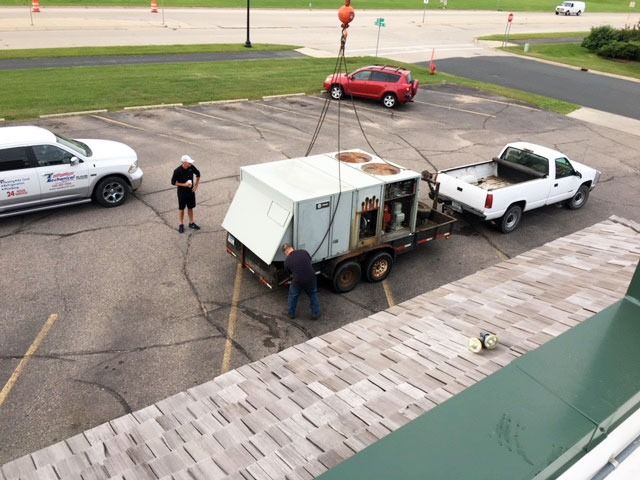 Workers load a white utility trailer onto a pickup truck in a parking lot, with another truck and car nearby.
