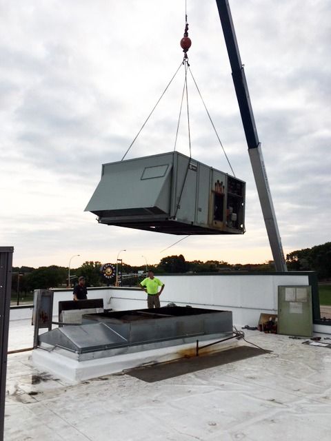 Crane lifting a gray modular unit onto a rooftop with workers nearby.