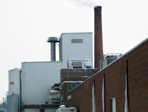 Industrial factory buildings with a tall brick smokestack beside a brick wall under an overcast sky