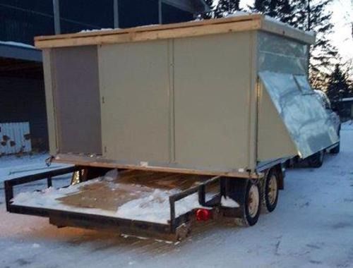 Small gray trailer or tiny house on a snowy lot, with a metal frame and covered side panel.