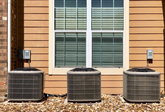 Three outdoor air conditioning units beneath a window on a wood-sided house.