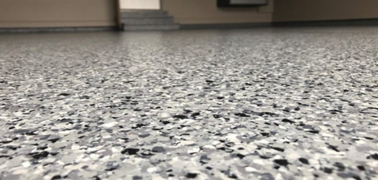 A close-up view of a gray, speckled epoxy floor, with walls and a set of stairs visible in the background.